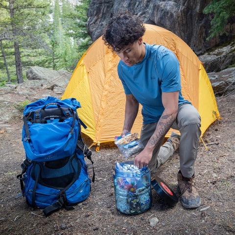Backpacker using BV475 Trek Portable Bear Resistant Canister near orange tent, showing mid-size design and rounded corners.
