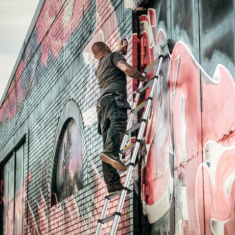 Man using a 12.5 Foot Telescoping Aluminum Ladder for Home DIY and Outdoor Use, painting graffiti on brick wall