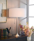 Stowe Table Lamp in Brushed Steel Finish With White Textured Drum Shade on a wooden nightstand near flowers in a living room setting.