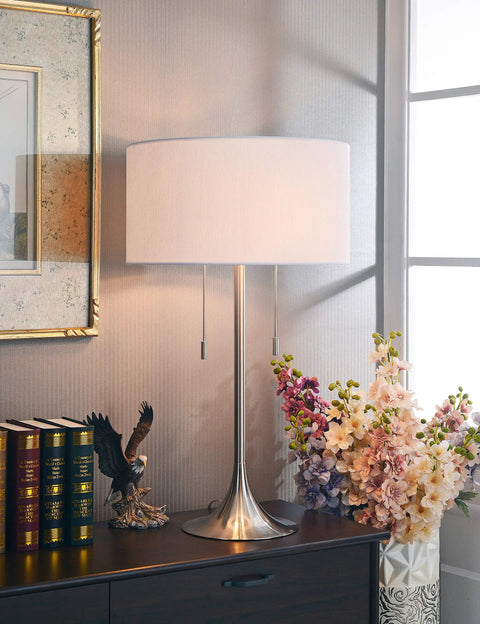 Stowe Table Lamp in Brushed Steel Finish With White Textured Drum Shade on a wooden nightstand near flowers in a living room setting.