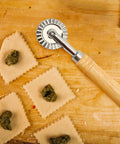Pizza cutter wheel with wooden handle and fluted zinc alloy blade, 16 cm long, next to square ravioli on a wooden board