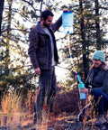 Man using Gravity Water Filter Portable Purifier with 3L bag and tree strap while woman collects water in forest setting