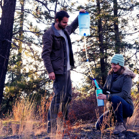Man using Gravity Water Filter Portable Purifier with 3L bag and tree strap while woman collects water in forest setting