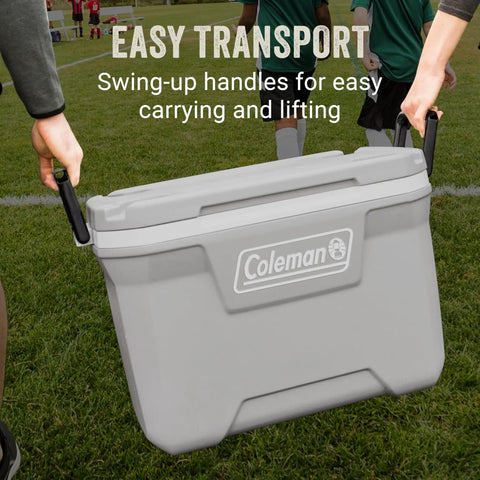 Two people carrying a Coleman cooler with swing-up handles on a field. Classic Series Insulated Portable Cooler With Leak Resistant Build.
