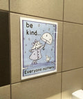 8.5x11 Clear Acrylic Sign Holders With No Drilling Mount, holding 'Be Kind' poster in restroom setting, wall mounted.