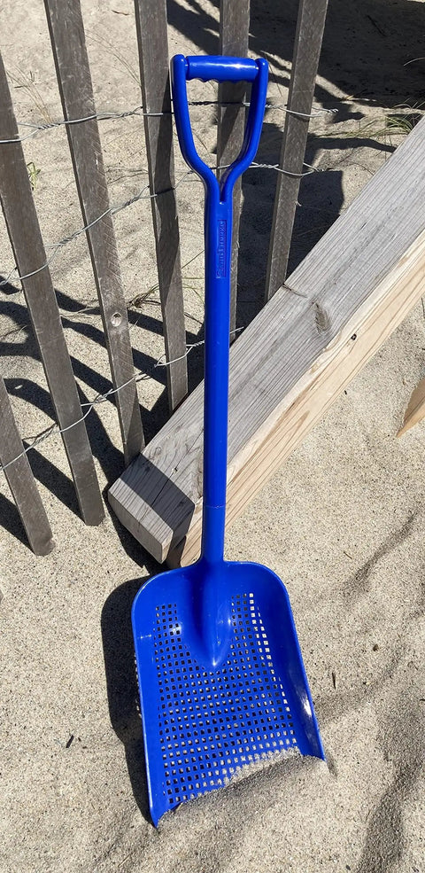 Sand Dipper Treasure Shovel Digger Sifter For Beach Cleanup And Barn Chores against a sandy beach fence.