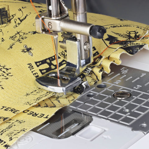 Front view of silver sewable shirring gathering presser foot on a low-shank sewing machine with yellow printed fabric.