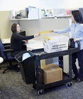 Two women exchanging mail at an office with a Top Tier Heavy Duty Two Shelf Utility Cart With Durable Structural Foam Lipped Trays.