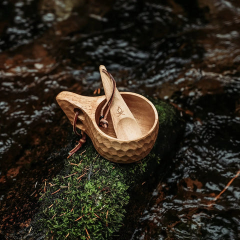 Kuksa Wooden Cup Handcarved 8 Oz Coffee Mug with Lanyard and Carabiner resting on moss by a river, durable hardwood design for outdoors.