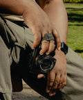 Person holding Micro Clutch L Plate Hand Strap for Mirrorless Cameras Low Profile Finger Loop with Battery Access ARCA Base