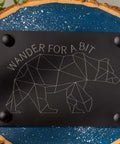 Front view of a black rectangular home decor sign with a geometric bear and Wander for a Bit text on a speckled surface.