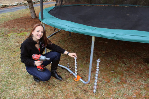 Woman installing Top Shelf 18 Inch Aluminum Screw Earth Anchor for trampoline; reusable, heat-treated, durable design.
