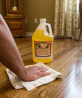 Orange Oil Wood Cleaner And Polish Spray For All Wood Surfaces Restorer And Conditioner Delivers Boss Level Shine On Cabinets Floors And Furniture Hardwood Floor Safe No Wax Build Up One Gallon bottle on wood floor with hand using cloth.