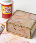 Glass box decorated with paper using All In One Decoupage Glue Sealer And Finish For Wood Paper Fabric Quick Dry Glossy Finish Non Toxic Craft Solution 16 Oz Made In USA Pack Of 1 Great For DIY Projects, with brush and bottle nearby.