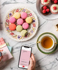 Hand using ColorReader Pro Digital Colorimeter And Paint Matching Tool for color matching macarons on a marble table with tea setup.