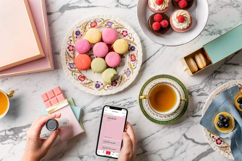 Hand using ColorReader Pro Digital Colorimeter And Paint Matching Tool for color matching macarons on a marble table with tea setup.