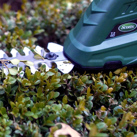Close-up of Cordless Grass Shear And Shrub Trimmer With Wheeled Extension Handle Seven Point Two Volt Lithium Ion Battery on a bush.