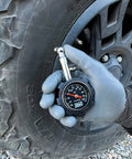 Front view of JACO Deluxe Tire Pressure Gauge, 100 PSI, held in a gloved hand beside a tire.