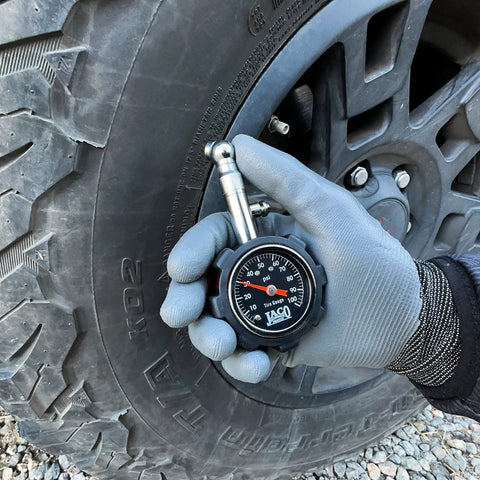 Front view of JACO Deluxe Tire Pressure Gauge, 100 PSI, held in a gloved hand beside a tire.