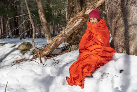 Person wrapped in Ninety Percent Heat Reflective Emergency Bivvy with Survival Whistle in snowy forest, ultra lightweight shelter