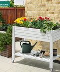 Elevated Garden Bed Kit With Legs And Wheels Large Resin Planter Box Stand With Drain Plug And Storage Shelf Perfect For Plants Vegetables Fruits Outdoor Durable Grayish White on patio filled with colorful flowers.