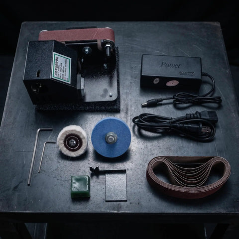Compact Mini Electric Belt Sander With A Kickass Motor For Handheld Wood Sanding And Fast Metal Polishing Quick Rust Removal And Smooth DIY Finishes Built Like A Rock Solid Workhorse displayed with accessories on table.