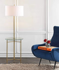 59 Inch Brass Gold Metal And Glass Side Table With Built In LED Floor Lamp Modern Transitional Iron Standing Lamp With White Linen Drum Shade UL Listed LED Bulb Included beside a blue chair.
