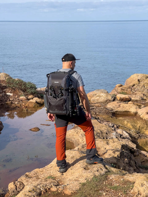 Person hiking with Alta Sky 62 Camera Backpack For 200 To 600mm Telephoto On DSLR Or Full Frame Mirrorless on rocky coast.