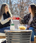 Two women enjoying drinks around a Tabletop Fire Pit Cover For Patio Pits 25.6 Inch Durable Stainless Steel Lid Weather Resistant Outdoor Fire Pit Protector Converts Fire Pit Into A Table For Chill Entertaining Gold.