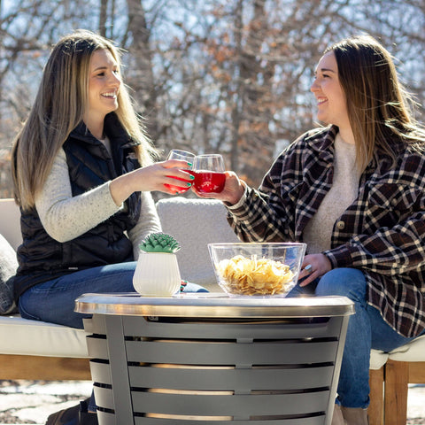 Two women enjoying drinks around a Tabletop Fire Pit Cover For Patio Pits 25.6 Inch Durable Stainless Steel Lid Weather Resistant Outdoor Fire Pit Protector Converts Fire Pit Into A Table For Chill Entertaining Gold.