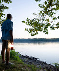 Person holding 30 Day 1000 Lumen LED Lantern With Glow In The Dark Power Button And Built In Hook for camping by a lake.