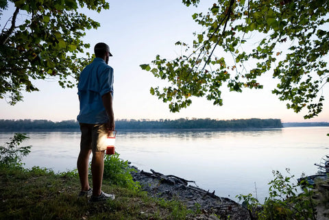 Person holding 30 Day 1000 Lumen LED Lantern With Glow In The Dark Power Button And Built In Hook for camping by a lake.