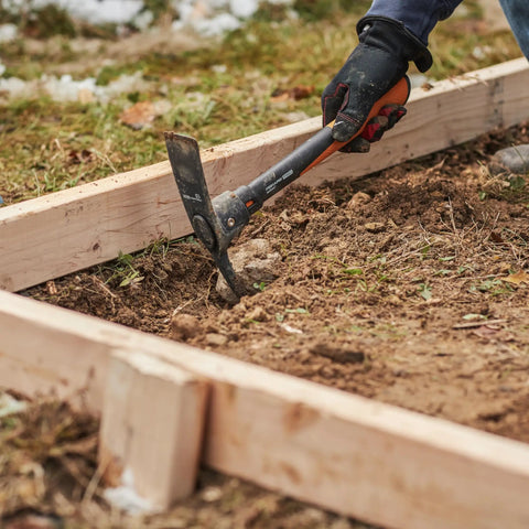 Using IsoCore Pickaxe Mattock With Shock Absorbing Handle Dual Head For Roots And Rocks 1.5 Lb Heavy Duty Tool on soil beside wood frame
