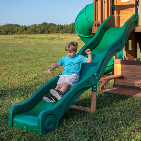 Child sliding down the wave slide of the Cedar Cove Wooden Swing Set with Spiral Tube Slide and Wave Slide, Elevated Clubhouse, in a backyard.