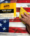 Pure Gold Superior Tack Cloths Pack Of 12 For Auto Body And Woodworking Prep For Slick Paint Jobs And Pristine Finishes 18 By 36 Inch Micro Apertured Dust Trapping Cloths That Grab Dust Lint And Sanding Particles, shown on a wooden flag surface with one being used by hand.