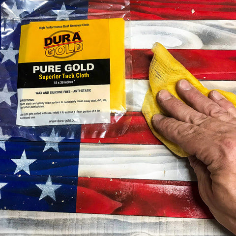 Pure Gold Superior Tack Cloths Pack Of 12 For Auto Body And Woodworking Prep For Slick Paint Jobs And Pristine Finishes 18 By 36 Inch Micro Apertured Dust Trapping Cloths That Grab Dust Lint And Sanding Particles, shown on a wooden flag surface with one being used by hand.