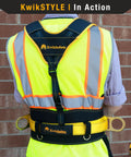 Back view of person wearing KwikSafety harness and reflective vest against brick wall.