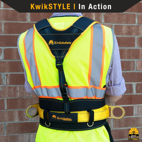 Back view of person wearing KwikSafety harness and reflective vest against brick wall.