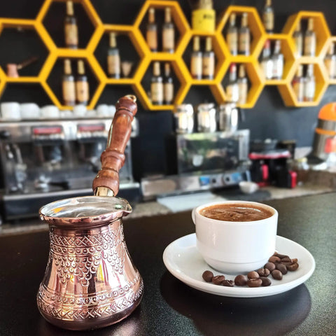 Top Notch Solid Copper Cezve for Turkish Greek Arabic Coffee with engraved finish and wooden handle next to coffee cup in cafe