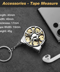 Compact keychain tape measure on a dark surface, showing dimensions and weight, beside a smartphone and a wireless earbud.