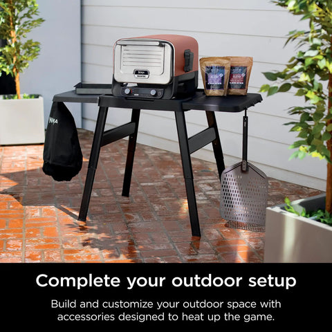 Outdoor setup with Woodfire Outdoor Perforated Pizza Peel, Lightweight Aluminum, Perforated Design to Shake Off Flour and Prevent Dough Sticking, Compatible With OO100 Series Outdoor Ovens, Gray and Black on table with pizza oven and accessories.