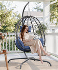 Woman reading on a Foldable Egg Chair With Stand For Indoor And Outdoor Use Wicker Rattan Swing Chair 485 Lbs Capacity Strong Iron Frame UV Resistant Cushion Patio Balcony Blue on a patio.