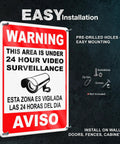The Real Deal 24 Hour Security Camera Alert Eight By Twelve Inch Reflective Aluminum Yard Sign bold bilingual lettering mounted on a wall.