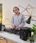 DJ using DM 50D BT 5 Inch Desktop Monitor System With Bluetooth in a modern studio setup with geometric wall art and greenery.