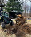 Compact tractor with Beefy Mini Wrecker Stump Bucket lifting large tree stump in yard, showcasing heavy duty leverage capability.