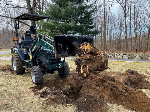 Compact tractor with Beefy Mini Wrecker Stump Bucket lifting large tree stump in yard, showcasing heavy duty leverage capability.