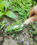 Hand holding large trowel from 5 Piece All In One Garden Tool Set With Large Trowel, Transplanter, Weeder, Hand Rake And Cultivator For Quick Digging, Planting And Weeding, Durable Steel Heads And Ergonomic Handles For Easy, Comfy Garden Work, digging in soil near plants.