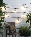 Outdoor LED String Lights 48 Feet With 16 Edison Style S14 2700K Soft White Bulbs On Black Cord For Patio Deck Or Backyard Vibe UL Listed Weatherproof And Long Lasting illuminating white brick wall with plants and chair