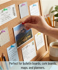 Hand placing red push pin on bulletin board surrounded by assorted notes and photos. Bulk Assorted Push Pins For Bulletin Boards Featuring Hella Bright Plastic Heads And Sharp Metal Tips That Gonna Keep Your Notes Secure And Organize Your Space Like A Pro.