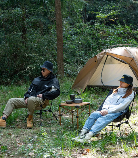 Two people sitting in Ultralight Camping Folding Beach Chair With Anti Sinking Large Feet And Back Support Webbing by a tent in the woods.